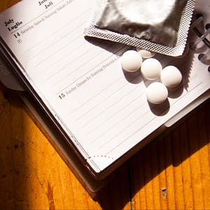 illustration for Sex Tips Series entitled Sex by schedule showing an open diary on a wooden desktop with a condom in silver foil and some round white pills 